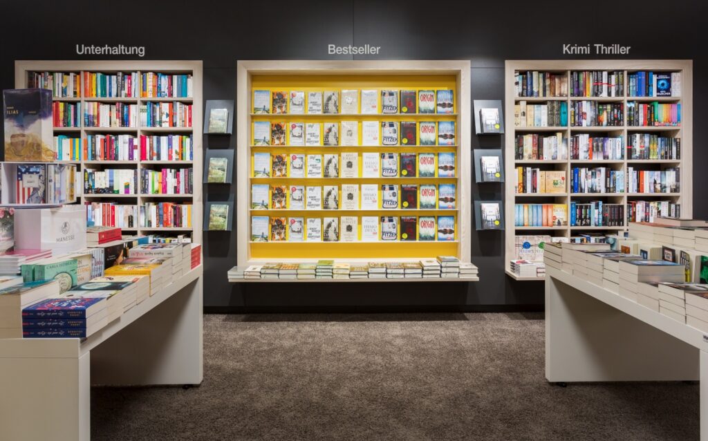 Verkaufsraum mit beleuchteten Genre-Wänden – Bücher, Teppich und gedämpfte Beleuchtung schaffen eine ruhige Beratungszone