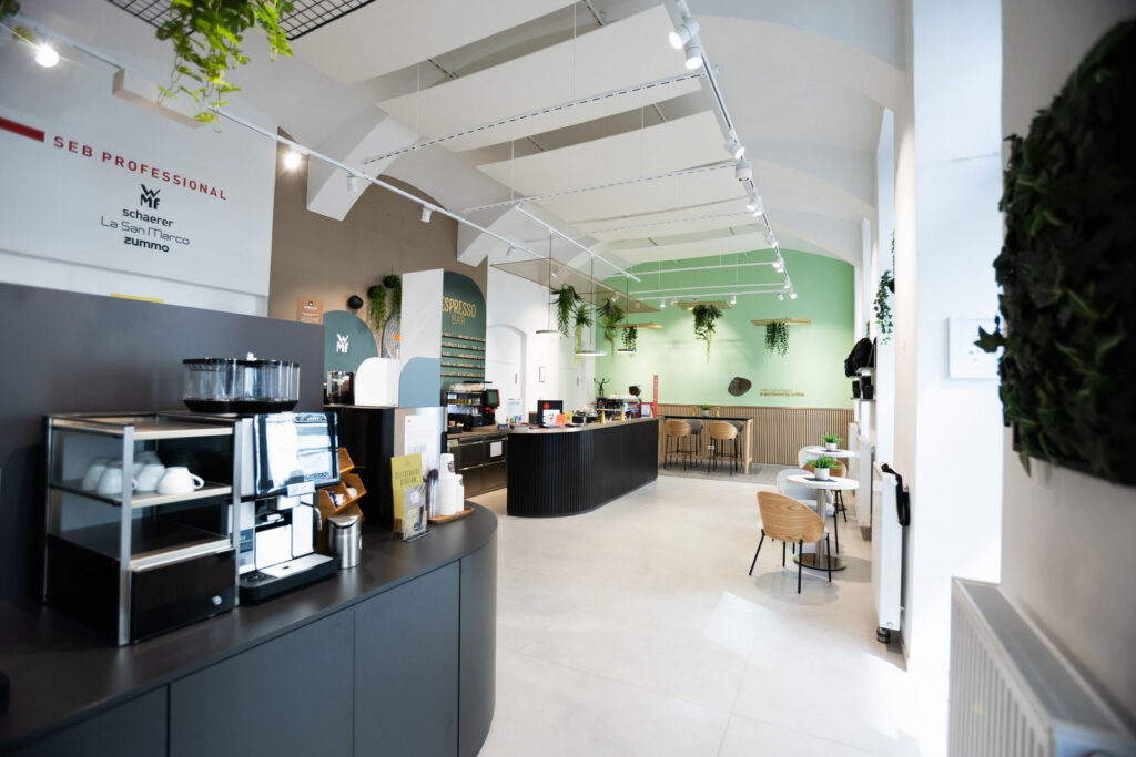 Bright, modern cafe interior with a black curved counter, espresso machines, and a lime-green wall; hanging plants and wooden chairs nearby.