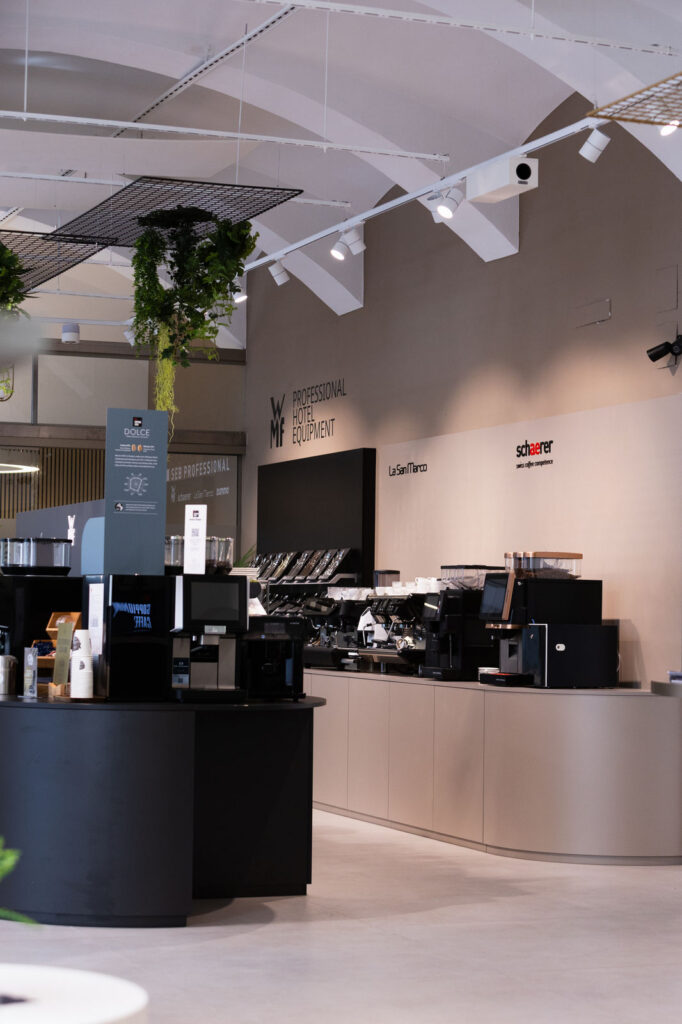 Modern cafe counter inside a showroom, with commercial espresso machines and equipment on display, plants overhead.