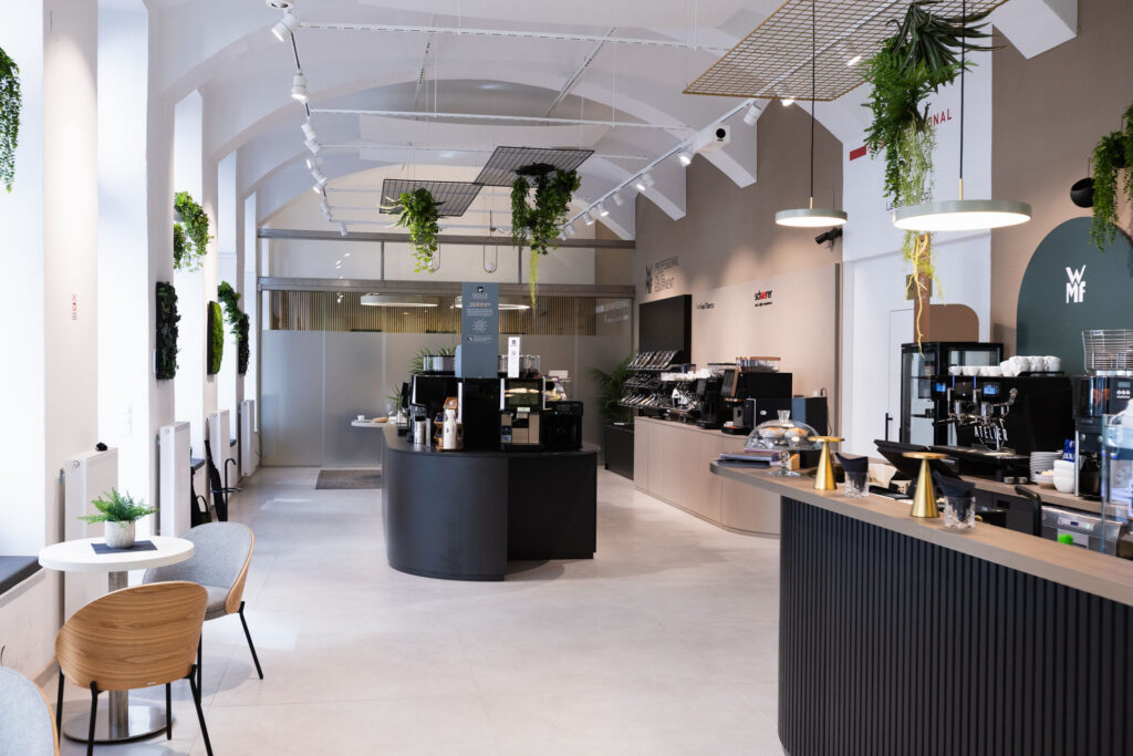 Bright cafe interior with a long dark curved counter, pastry display, and hanging plants in an airy space.