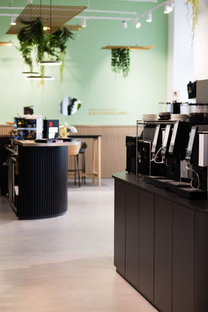 Modern cafe counter with multiple espresso machines on the right and hanging plants above a green wall.