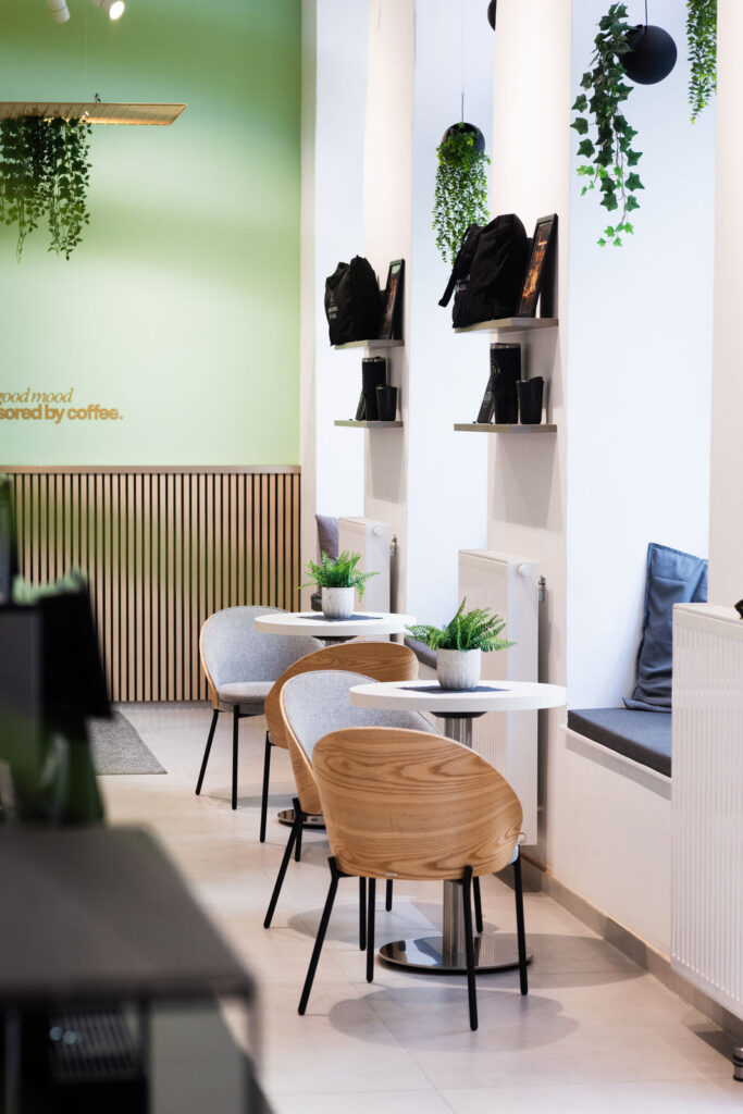 Café seating area with round white tables, wooden chairs, gray cushions, green wall, and hanging plants.