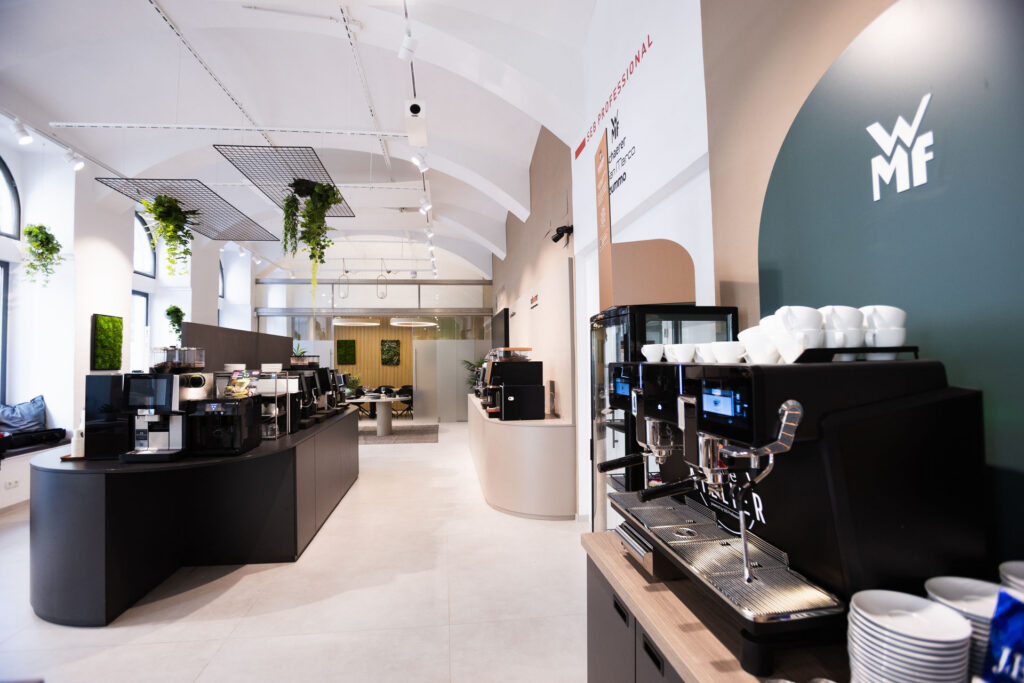 Bright, modern cafe interior with a row of espresso machines along a curved counter and hanging greenery overhead.