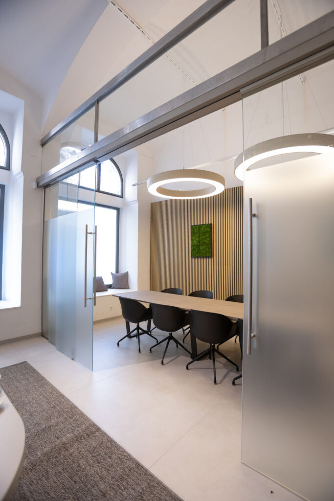 Modern conference room with a long wooden table, six black chairs, and circular pendant lights; glass partition and wooden slat wall behind. Readable windows provide natural light.
