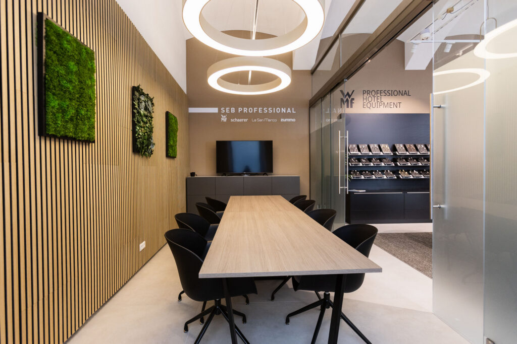 Modern conference room with a long wood table, black chairs, circular pendant lights, and a glass wall displaying a hotel equipment showroom.