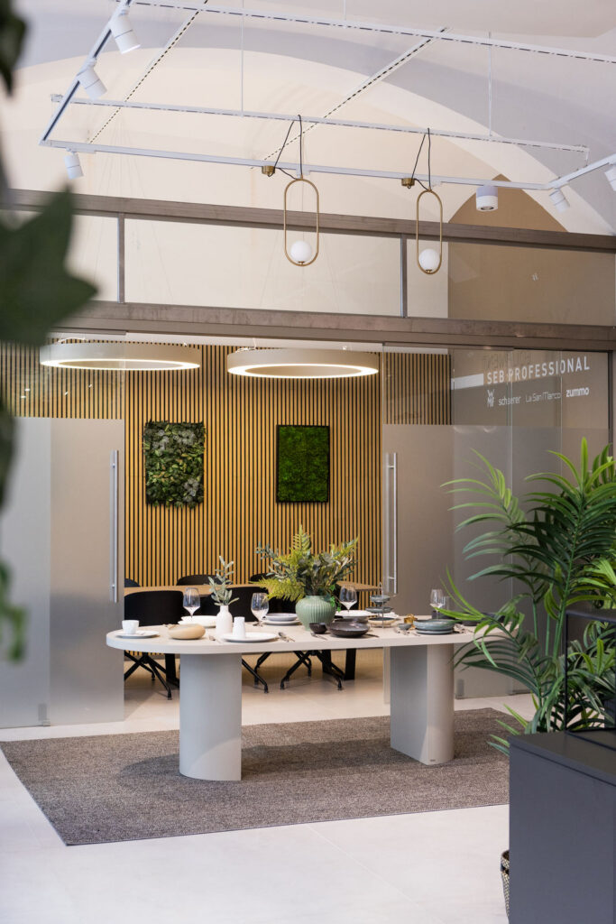 Modern office dining area with a long white table set for a meal, plants, and wood slat wall behind.